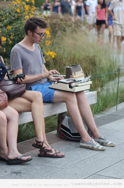 Chico hipster sentado en el banco de un parque con una máquina de escribir en el regazo