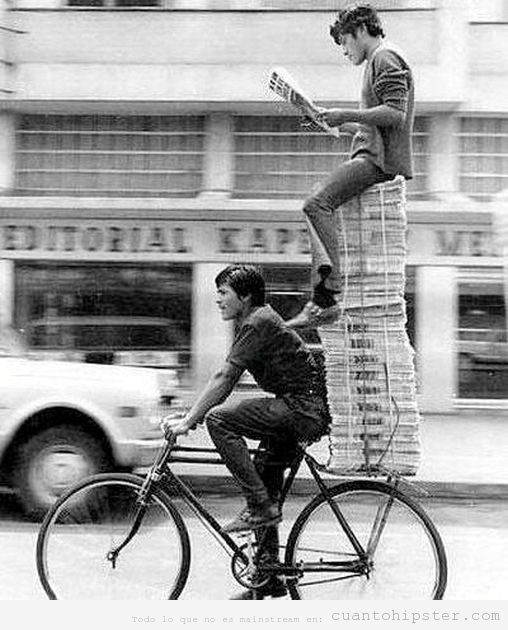 Foto antigua de un repartidor de periódicos en bicicleta y uno sentado encima de la pila
